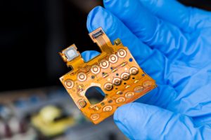 Round button switches, LED diodes or flex flat cable with connector on bent plastic board on dark blur background. E-waste disposal